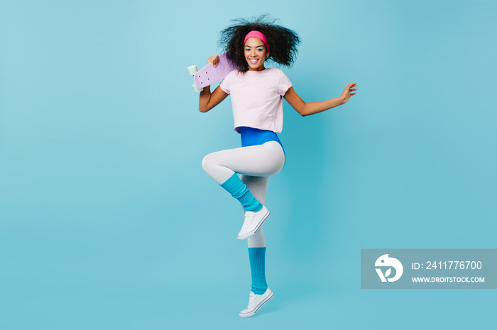 Good-humoured fitness girl jumping on blue backgorund. Studio shot of glad african woman dancing with purple skateboard.