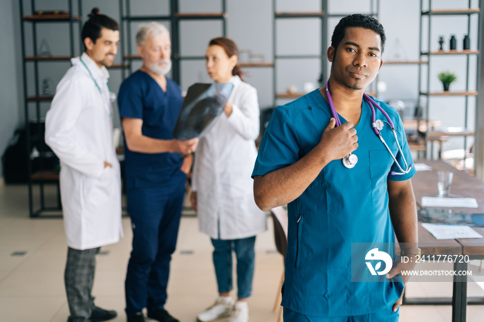 Portrait of confident African male surgeon doctor in blue uniform standing in medical meeting office, looking at camera. Multiethnic team of physician working on background, discussing scan of patient