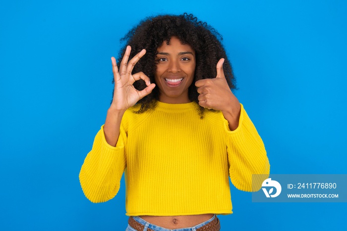 young woman with afro hairstyle wearing orange crop top over blue wall  feeling happy, amazed, satisfied and surprised, showing okay and thumbs up gestures, smiling