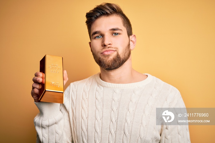 Young blond man with beard and blue eyes holding gold ingot over isolated yellow background with a confident expression on smart face thinking serious