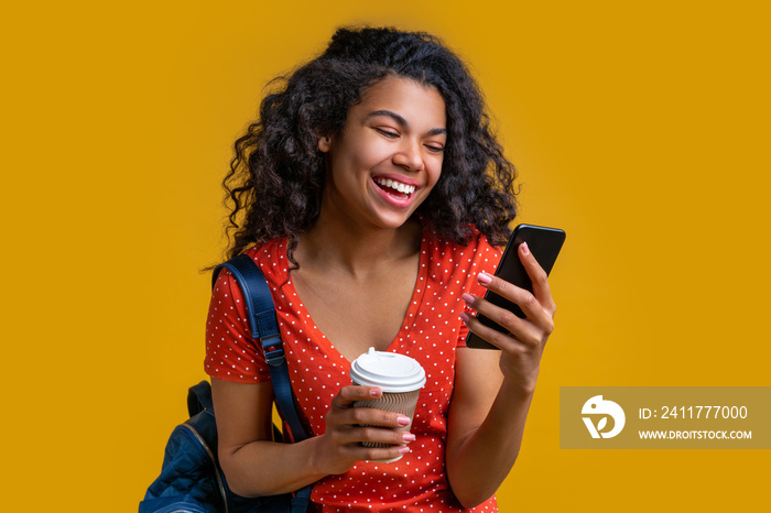 Close up of beautiful smiling dark skinned girl with a backpack holding paper cup of coffe in hands and reading message on her mobile phone