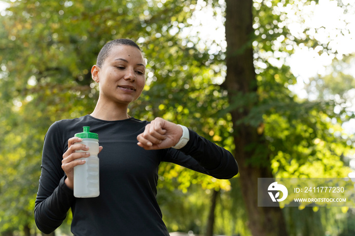 Athletic woman holding water bottle and checking smart watch in park