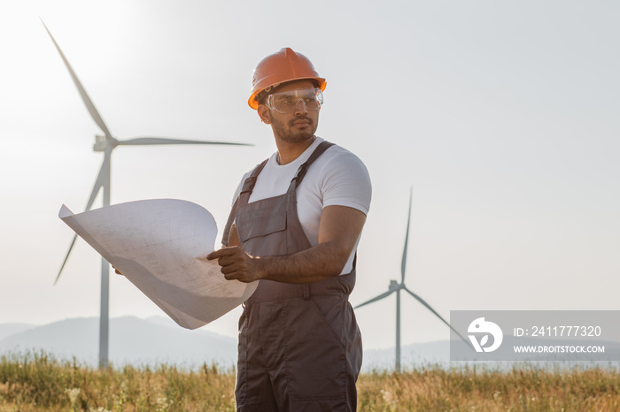 Indian technician in overalls, helmet and glasses checking blueprints of windmills while standing at rural area. Competent industrial worker controlling process of green energy production.