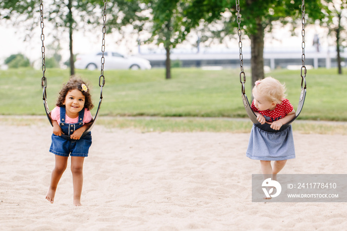 Portrait of two happy smiling little toddlers girls friends swinging on swings at playground outside on summer day. Happy childhood lifestyle concept. Toned with film pastel faded filters colors.