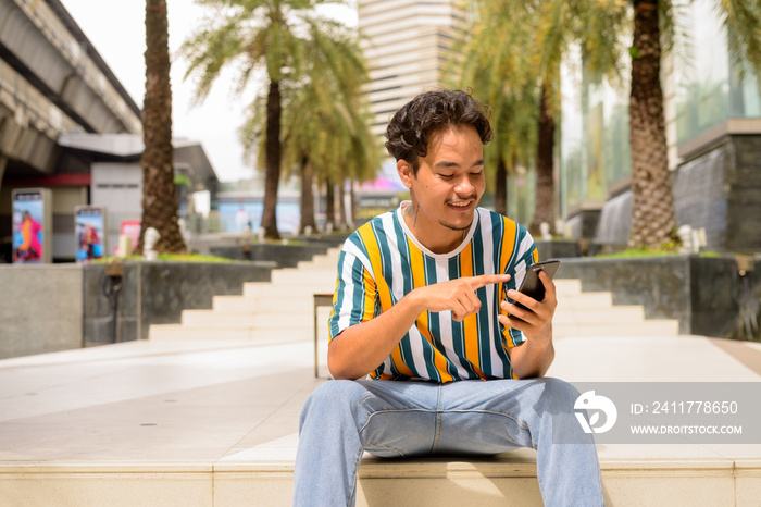 Portrait of happy multi-ethnic young man wearing colorful shirt outdoors during summer while using mobile phone