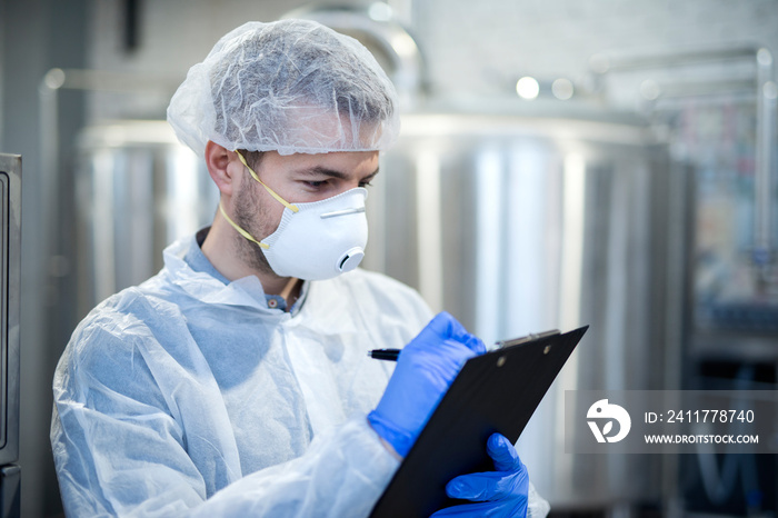 Technologist in white suit wearing hairnet and protective mask checking production in industrial factory.