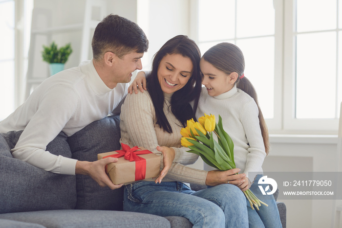 Happy mother’s day. Father and baby daughter congratulates mom with flowers and a postcard in a room