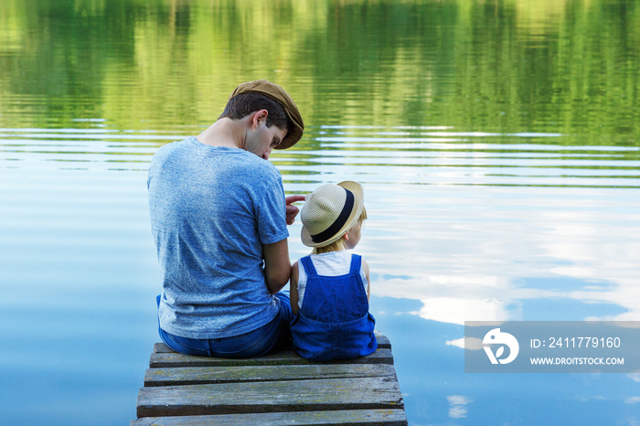 Dad and a tiny girl in dungarees relax at the river. Back view, copy space.