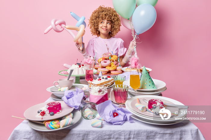 Horizontal shot of thoughtful curly haired holds bunch of inflated balloons wears festive dress and gloves stands near table with full of desserts celebrates birthday isolated over pink background