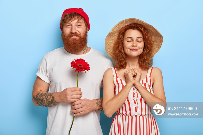 People, closeness, love concept. Handsome bearded red haired boyfriend has date with sweetheart, holds gerbera, knows women like flowers. Pleased lady in summer hat, dress spends free time with lover