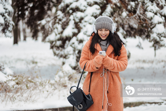Pretty young long-haired woman in pink coat enjoy winter day and listening music with headphones at snowy park