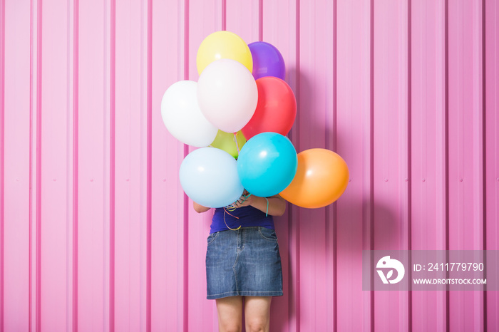 Girl holding multicolored helium balloons against pastel pink background.