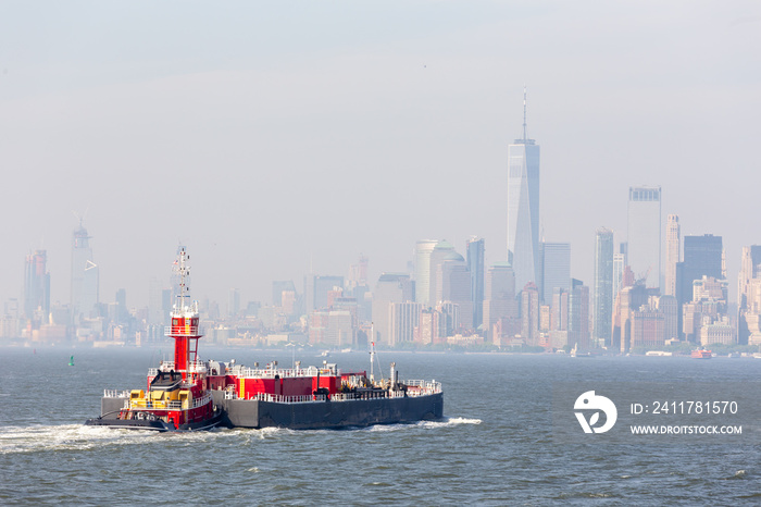 Freight tug pushing cargo ship to the port in New York City and Lower Manhattan skyscarpers skyline in background. New York City, USA.