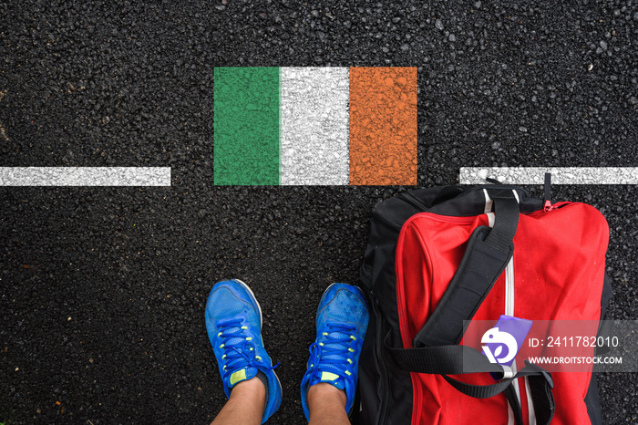 a man with a shoes and travel bag is standing on asphalt next to flag of Ireland and border