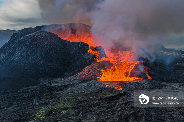 Daytime volcanic eruption on Reykjanes peninsula. Side view of the crater opening with flowing lava. Crater from Fagradalsfjall volcano in Iceland in GeoPark. Clouds and steam in the sky