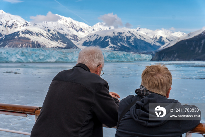 Old couple on a cruise ship in Alaska. Husband and wife are enjoying an amazing view of Hubbard Glacier and iceberg. Snow peaks and ice all around in the sea.