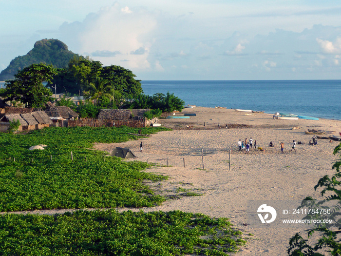 Fin de journée sur la plage d’un village africain des Comores. Les barques de pêche sont renversées sur le sable, quelques personnes discutent, à l’arrière-plan les premières maisons du village