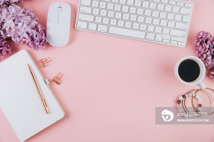 Spring flat lay top view home office workspace - modern keyboard, cup of coffee and notebook with lilac flowers on a pink desk background