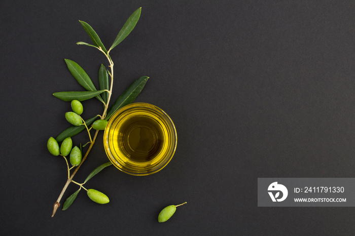 Top view of olive oil in a glass bowl and branch with green olives on the black background. Copy space.