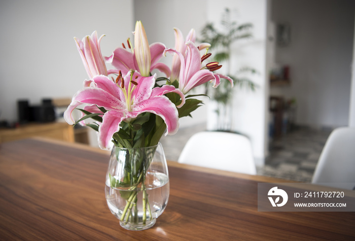 Bouquet pink lily flowers in glass vase on wood table in room.