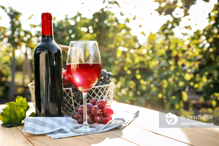 Glass and bottle of red wine with fresh grapes on wooden table in vineyard