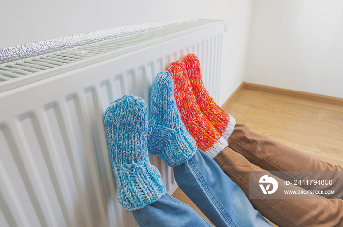 Family wearing colorful pair of woolly socks warming cold feet in front of heating radiator in winter time. Electric or gas heater at home. The symbolic image of the heating season at home.