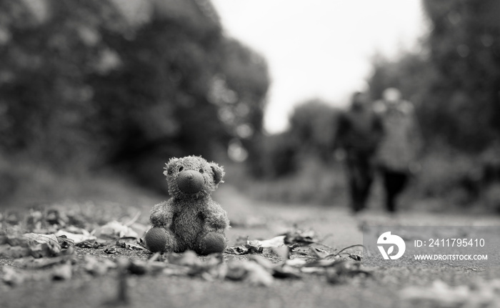 Lost Teddy bear with sad face sitting on footpath with blurry people Lonely bear doll sit down on the brick floor in gloomy day, Lost toy or Loneliness concept, International missing Children