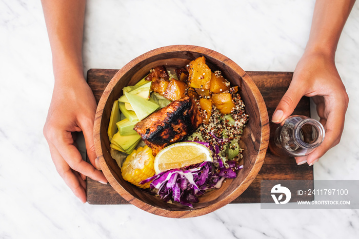 Woman eating meal with fried salmon fish steak, quinoa, avocado, corn, cabbage salad and baked pumpkin in wooden bowl. Healthy organic food concept. White marble table surface.