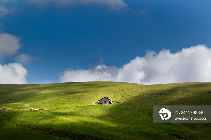 Grassy hill with an isolated cabin in the center, blue sky with clouds. Play of light on the Piani dell’Avaro, Val Brembana, Bergamo, Italy. Mountain landscape on a summer day. Minimal composition