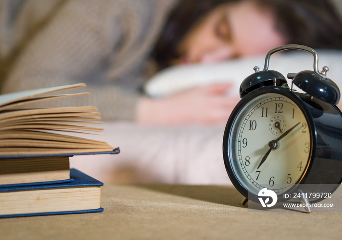 A black alarm clock on a bedside table next to a pile of books. Blurred in the background, a woman sleeps on a bed.