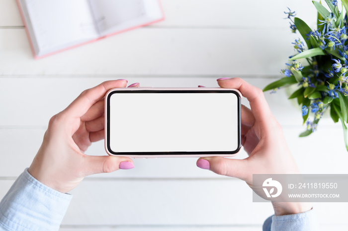 Female hands with a smartphone. White blank screen. Table with notebook and flowers on background