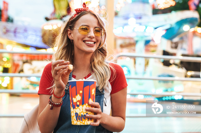 Positive young blonde woman in amusement park eat popcorn.