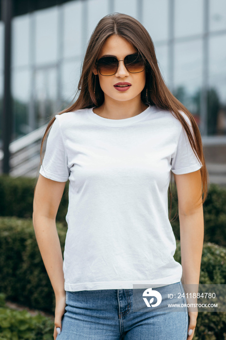 Stylish brunette girl wearing bwhite t-shirt and glasses posing against street , urban clothing styl