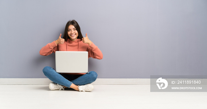 年轻的学生女孩拿着笔记本电脑在地板上竖起大拇指