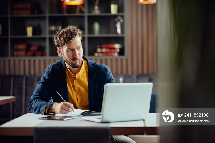 Young serious bearded Caucasian blogger dressed smart casual writing notes in agenda and looking at 