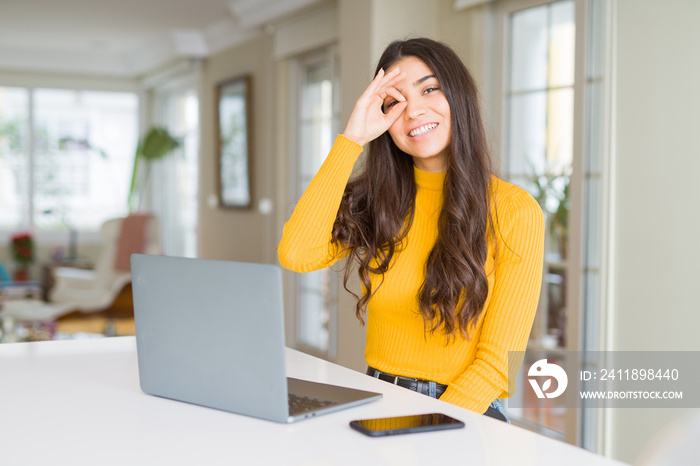 Young woman using computer laptop doing ok gesture with hand smiling, eye looking through fingers wi