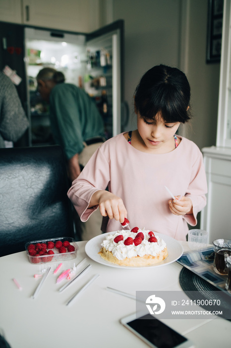 Girl decorating cake with strawberries at table in kitchen