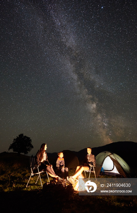 年轻的家庭旅行者在夏季露营时一起坐在椅子上，在篝火和游客旁边