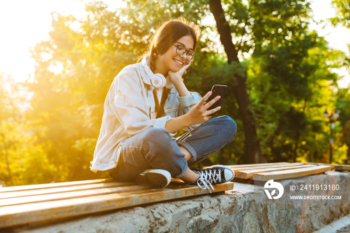 Happy cute young student girl wearing eyeglasses sitting outdoors in nature park using mobile phone.