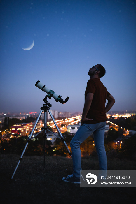 Astronomer with a telescope watching at the stars and Moon with blurred city lights in the backgroun