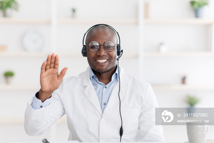 Friendly african american doctor in headphones waving hand to camera, sitting at table in office