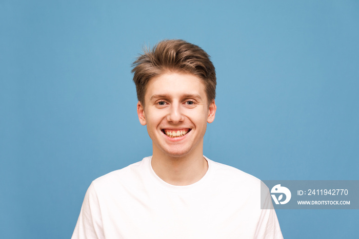 Smiling young man in a white T-shirt smiles and looks at the camera, a close portrait on a blue back