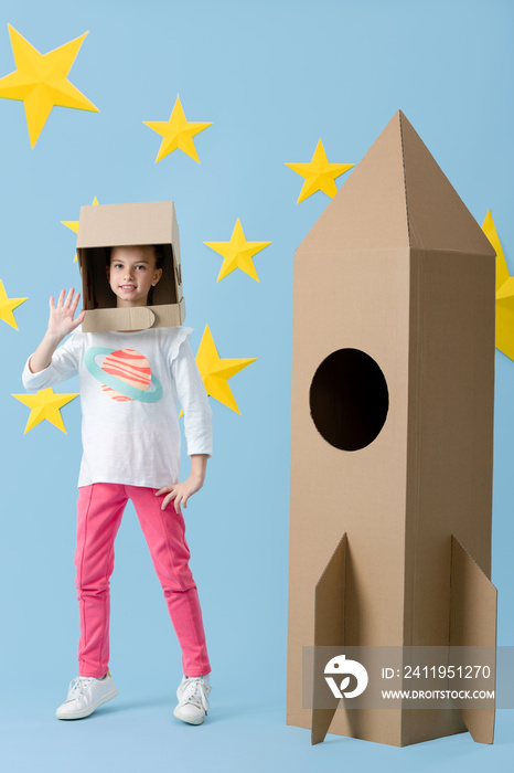戴着头盔的快乐孩子站在纸板火箭附近，在蓝色背景上与星星挥手