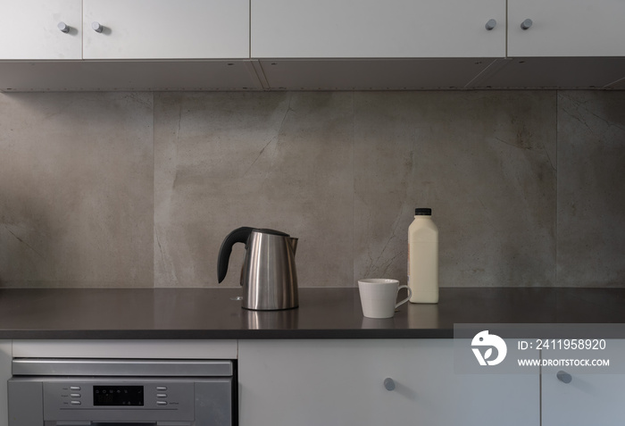 Stainless steel kettle with white cup and milk bottle on dark grey kitchen bench (selective focus)