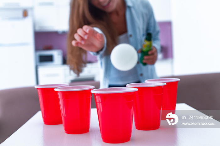 Woman with drinks having fun and enjoying beer pong game on table at home.