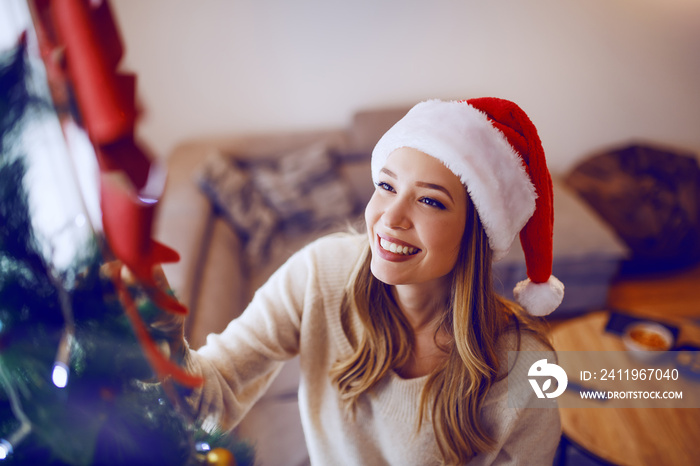 头戴圣诞老人帽，笑容灿烂的高加索金发女子站在客厅里装饰