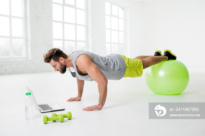 Man watching online trainig with laptop training on the fitball in the gym