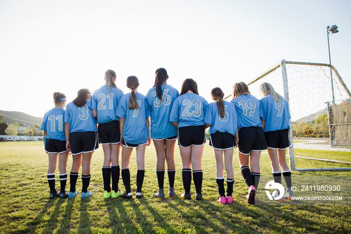 Rear view of girls wearing soccer uniforms standing on grassy field against sky