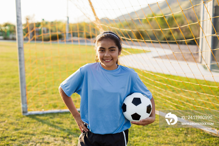 Portrait of smiling soccer player with hand on hip holding ball while standing against net