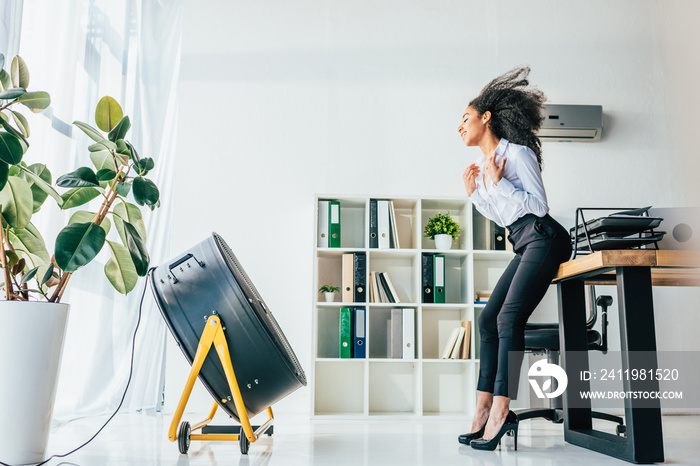 smiling african american businesswoman standing in front of blowing electric ventilator in office